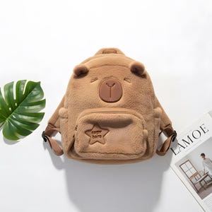 May include: A brown plush backpack shaped like a bear, featuring bear ears, a face, and a small front pocket with a star and the text "copy bara". The backpack has adjustable straps and a top handle.