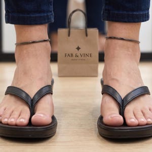 Op de afbeelding: Zwarte teenslippers met een dikke zool worden gedragen. Een bruine papieren boodschappentas met de tekst "FAB & VINE" staat op de achtergrond. De persoon draagt een donkerblauwe jeans en een zilveren enkelbandje.