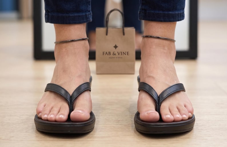 Op de afbeelding: Zwarte teenslippers met een dikke zool worden gedragen. Een bruine papieren boodschappentas met de tekst "FAB & VINE" staat op de achtergrond. De persoon draagt een donkerblauwe jeans en een zilveren enkelbandje.