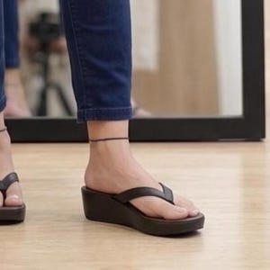 Op de afbeelding: Bruine leren sleehak sandalen met een teenslipper ontwerp. De sandalen hebben een plateauzool en worden gedragen met een donkerblauwe jeans. Een bruine papieren boodschappentas is zichtbaar.