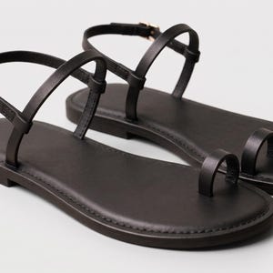 May include: A pair of black flat sandals featuring a toe loop design, thin ankle straps with small gold-toned metal buckles, and a smooth, rounded sole. The sandals have a minimalist construction with slender straps that secure around the ankle.