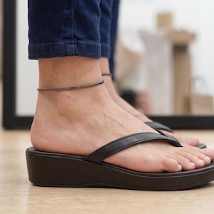 Op de afbeelding: Zwarte leren teenslippers met een donkerbruine sleehakzool. Een zilverkleurige enkelband is zichtbaar. De persoon draagt een blauwe spijkerbroek. De foto is binnenshuis gemaakt.