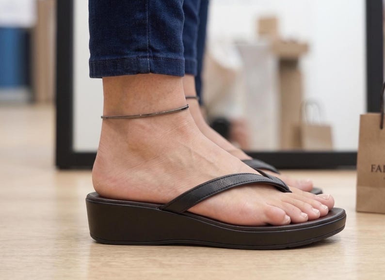 Op de afbeelding: Zwarte leren teenslippers met een donkerbruine sleehakzool. Een zilverkleurige enkelband is zichtbaar. De persoon draagt een blauwe spijkerbroek. De foto is binnenshuis gemaakt.