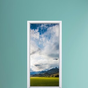 May include: A white door with a door handle opens to reveal a scenic view of a green field, mountains, and a cloudy blue sky.