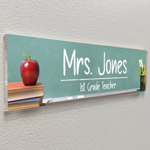 May include: A green chalkboard with a red apple, books, and pencils. The text on the chalkboard reads "Mrs. Jones 1st Grade Teacher".