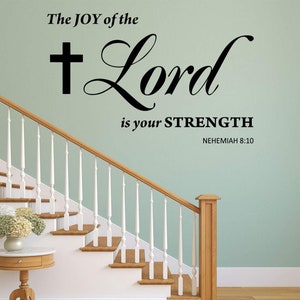 Peut inclure: Un autocollant mural avec la citation "The JOY of the Lord is your STRENGTH" avec une croix et le verset Néhémie 8:10. L'autocollant est sur un mur vert clair au-dessus d'un escalier en bois.