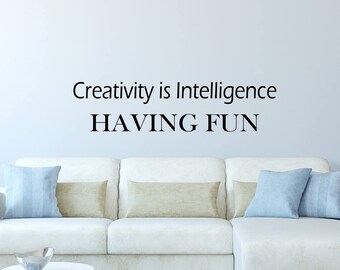 May include: A white sectional sofa with beige and blue throw pillows in front of a light blue wall with the quote "Creativity is Intelligence HAVING FUN" written in black.