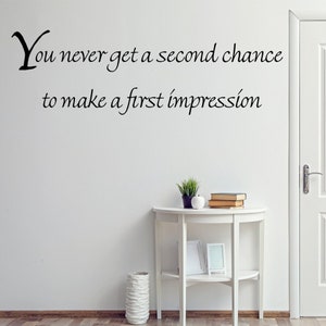 May include: White wall with a black vinyl decal that reads "You never get a second chance to make a first impression". A white table with a plant, books, and a picture frame sits in front of a white door.