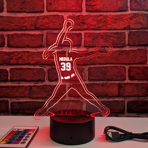 May include: A red LED night light shaped like a baseball player in a pitching pose. The player is wearing a jersey with the name "MEDULA" and the number "39" on the front. The night light has a black base and comes with a remote control and a USB charging cable.