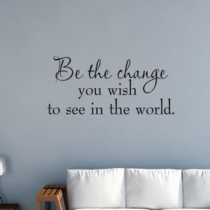 Op de afbeelding: Een muursticker met de quote "Wees de verandering die je in de wereld wilt zien." De sticker zit op een lichtblauwe muur boven een witte bank met houten poten.