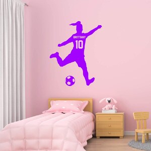 May include: Purple silhouette of a soccer player kicking a ball with the name "Brittany" and the number "10" on the jersey. The decal is on a pink wall in a bedroom.