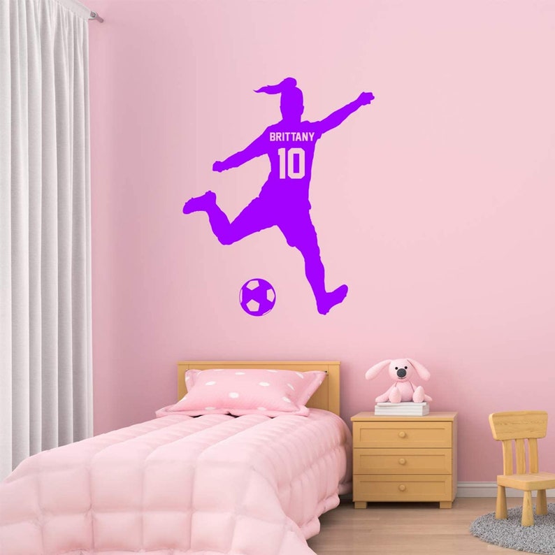 May include: Purple silhouette of a soccer player kicking a ball with the name "Brittany" and the number "10" on the jersey. The decal is on a pink wall in a bedroom.