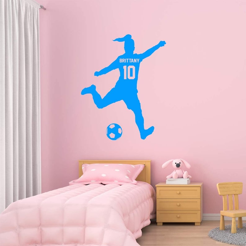 May include: A blue silhouette of a soccer player kicking a ball with the name "Brittany" and the number "10" on the jersey. The decal is on a pink wall in a bedroom with a bed, nightstand, and chair.