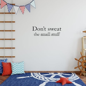 May include: A white wall with a black vinyl decal that reads "Don't sweat the small stuff". The wall is decorated with a string of red, white, and blue pennant flags. A blue rug with a white anchor design is on the floor.