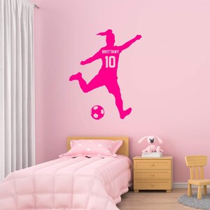 May include: Pink silhouette wall decal of a female soccer player kicking a soccer ball. The decal includes the name "Brittany" and the number "10".