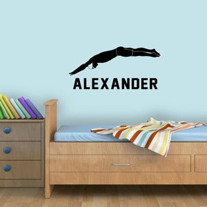 May include: A black silhouette of a person diving into a pool with the name "ALEXANDER" written below it. The image is on a light blue wall above a wooden bed frame with a striped blanket.