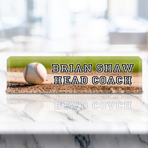 May include: A green and brown baseball field with a white baseball in the foreground. The text "BRIAN SHAW HEAD COACH" is printed in black on the image.