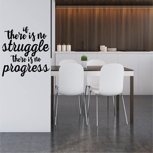 May include: A black and white inspirational quote on a white wall reads "if there is no struggle there is no progress". The quote is in a modern calligraphy style. The wall is in a dining room with a white table and chairs.