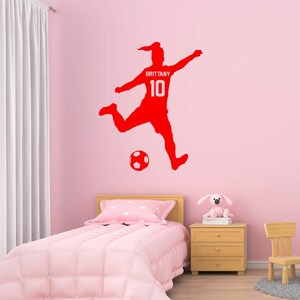 May include: Red silhouette of a soccer player kicking a ball with the name "Brittany" and the number "10" on the jersey. The decal is on a pink wall in a bedroom.