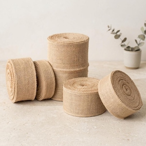 May include: Several rolls of natural burlap ribbon are arranged on a light-colored surface. The rolls vary in size and are made of a woven, tan-colored material. Some rolls are stacked, while others are unrolled, showcasing the ribbon's texture.