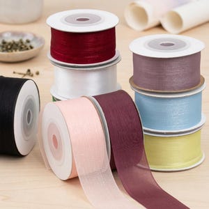 Pode incluir: Várias bobinas de fita decorativa em tons de vermelho, branco, preto, rosa, roxo, azul e amarelo. As fitas são feitas de um material transparente e texturizado e são exibidas em uma superfície de madeira clara. Ideal para artesanato e embrulho de presentes.