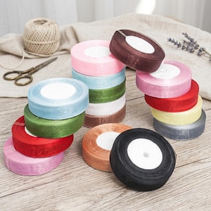 Puede incluir: Una colección de carretes de cinta de raso de colores en varios tonos, incluyendo rosa, azul, verde, rojo y marrón. Los carretes están apilados y dispuestos sobre una superficie de madera, con unas tijeras y cuerda en el fondo. Ideal para manualidades y envolver regalos.