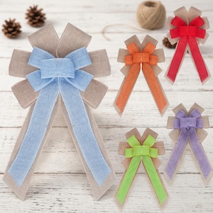 Könnte beinhalten: Eine Auswahl an dekorativen Schleifen in verschiedenen Farben. Die Schleifen haben ein mehrschichtiges Design mit einer Basis aus Jute und farbigen Bändern. Zu den Farben gehören Blau, Orange, Rot, Grün und Lila. Die Schleifen sind auf einer hellen Holzoberfläche angeordnet.