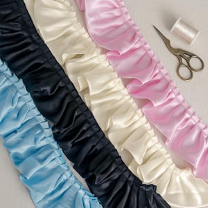 Könnte beinhalten: Nahaufnahme von mehreren gerüschten Satinbändern in verschiedenen Farben: Hellblau, Schwarz, Creme und Rosa. Eine kleine goldene Schere und eine Garnrolle befinden sich auf der rechten Seite der Bänder. Die Bänder sind für Näh- und Bastelarbeiten geeignet.