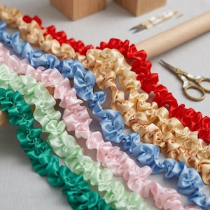 May include: A selection of ruffled satin ribbon in various colors, including red, gold, blue, pink, and green. The ribbons are arranged in a diagonal pattern, with a wooden dowel and small scissors in the background. Ideal for crafting and sewing projects.
