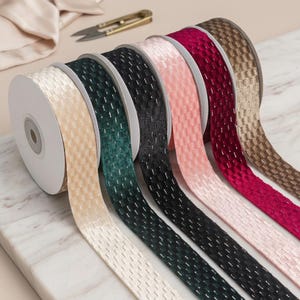 May include: A selection of decorative ribbon spools in shades of cream, beige, dark green, black, pink, burgundy, and brown. The ribbon features a textured weave with a metallic thread. Gold scissors and fabric are visible in the background.