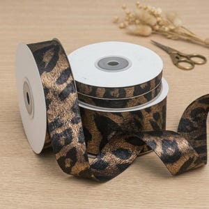 May include: Spools of decorative ribbon with a leopard print design in black and gold. The ribbon is on white spools, with a portion unrolled, revealing the pattern. Scissors and dried flowers are in the background.