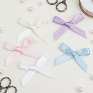 May include: An assortment of small satin bows in pastel colors: white, pink, lavender, and light blue. The bows are arranged on a white surface, with scattered pearls and a pair of small scissors. A roll of ribbon is visible in the background.