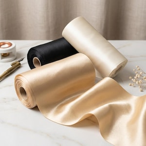 Puede incluir: Tres rollos de cinta de raso en negro, crema y dorado. La cinta dorada está desenrollada, mostrando su textura brillante. Los rollos están sobre una superficie de mármol blanco, con una pluma y un tintero en el fondo. Ideal para manualidades y envolver regalos.