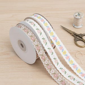 Könnte beinhalten: Drei Rollen Blumenband in Pastellfarben. Das Band zeigt rosa, gelbe und blaue Blumen. Eine Schere und eine Garnrolle sind ebenfalls auf einer Holzoberfläche zu sehen.