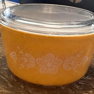 May include: A round, golden-yellow casserole dish with integrated side handles and a clear glass lid. The exterior of the dish features a white, repeating floral and leaf pattern encircling the center, providing a decorative contrast to the solid color.