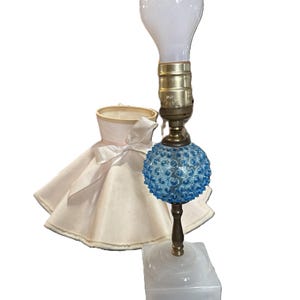 May include: A vintage table lamp with a white bulb, brass fixture, and a blue glass orb base. The lamp sits on a white square base. A white lampshade with a pink ribbon is next to the lamp.