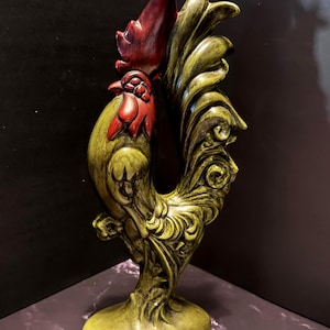 May include: A decorative rooster figurine featuring a textured olive green body and a deep red comb and wattle. The sculpture stands on a rounded base and displays intricate, swirling feather details along the tail and neck area.