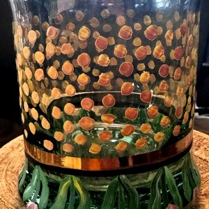 May include: A clear glass vessel featuring a hand-painted pattern of small orange and brown dots across the upper section. A horizontal metallic copper band encircles the middle, while the base displays green leaves and small pink flower buds.