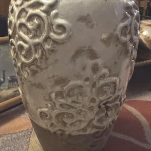 May include: A decorative ceramic vase with a cream-colored finish and embossed floral patterns. The vase has a wide, rounded body and a narrower neck. The base is a darker, earthy tone. The vase is approximately 12 inches tall.