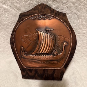 May include: A copper-colored Viking ship relief sculpture mounted on a dark wooden plaque. The ship has a large sail with a sun design, and is sailing on a wavy sea. The plaque has a decorative rope border.