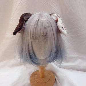 May include: A pair of plush animal ears, one brown and the other white with black spots, attached to a wig with gray and blue hair. The wig is displayed on a wooden stand against a white backdrop.