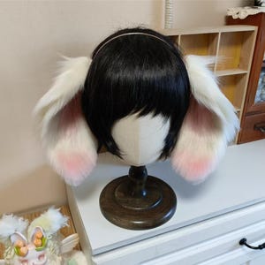Può includere: Un paio di orecchie da coniglio bianche e rosa attaccate a una parrucca nera su una testa di manichino. Le orecchie sono soffici con dettagli rosa all'interno. La testa del manichino è montata su un supporto di legno scuro.