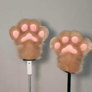 May include: Two plush paw-shaped hand covers. The paws are light brown with pink pads and claws. The furry paws are mounted on a white and black pole, likely for display or use as props. The background is a plain gray.
