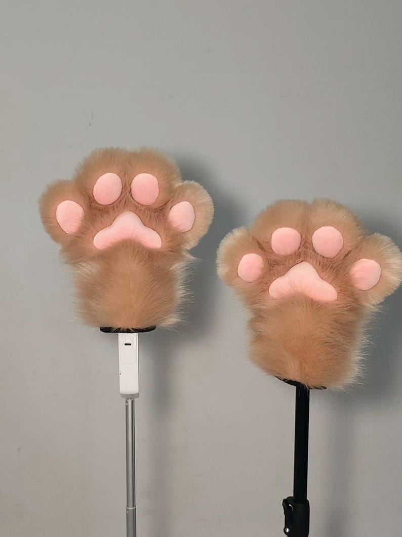 May include: Two plush paw-shaped hand covers. The paws are light brown with pink pads and claws. The furry paws are mounted on a white and black pole, likely for display or use as props. The background is a plain gray.