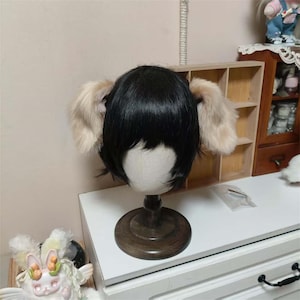 May include: A black wig with bangs and attached fluffy, light tan ears. The wig is displayed on a wooden mannequin head stand. The ears are a soft, textured material. The wig is likely for cosplay or costume use.