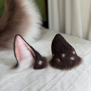 Puede incluir: Un par de orejas de animal de piel sintética en color marrón oscuro con manchas blancas. Una oreja está colocada para mostrar el interior, con pelo rosa claro y blanco, mientras que la otra muestra el exterior marrón oscuro con manchas.