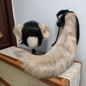 May include: A cosplay set featuring a black wig with attached light tan ears and a matching fluffy tail with a black ribbon. The tail and ears are made of faux fur. The items are displayed on a wooden cabinet.