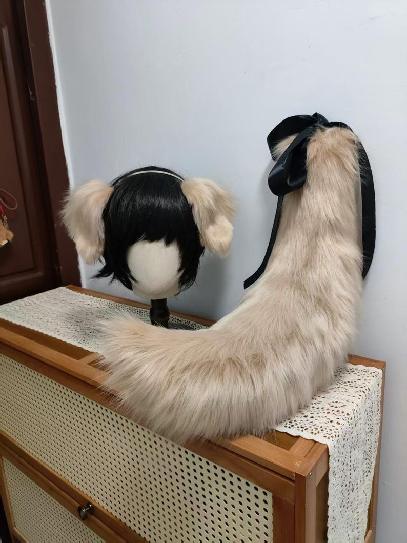 May include: A cosplay set featuring a black wig with attached light tan ears and a matching fluffy tail with a black ribbon. The tail and ears are made of faux fur. The items are displayed on a wooden cabinet.
