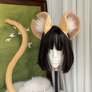 May include: A set of tan and cream faux fur animal ears attached to a headband, paired with a long, matching tan faux fur tail. The ears and tail are displayed on a mannequin head featuring a short black bob wig with white highlights.