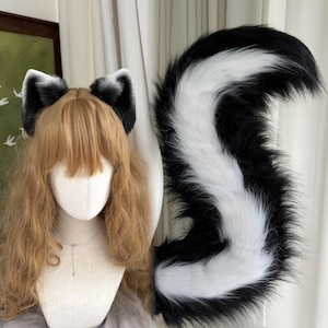 May include: A set of faux fur animal ears and a long, curved tail. The ears are black with white tips and inner details. The tail is long, fluffy, and features a prominent white stripe running down the center of the black fur, mimicking a skunk.